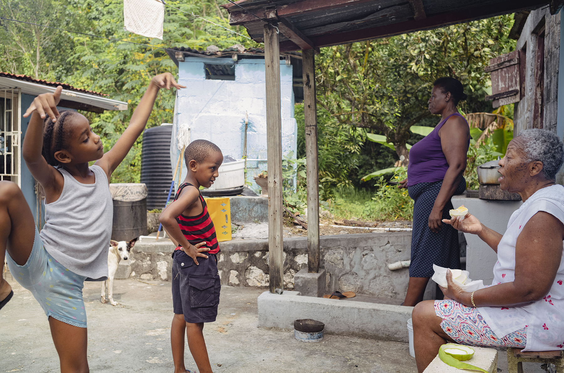 Photograph of one child dancing whilst another speaks with an older person.  Another person and a dog stood in the background.