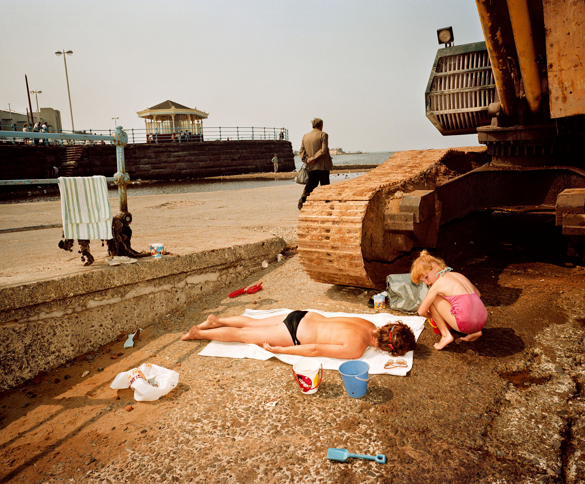 Photographer's image showing New Brighton seafront in 1983-85. A person laying on a beach towel with an excavator track poised to run over the person.