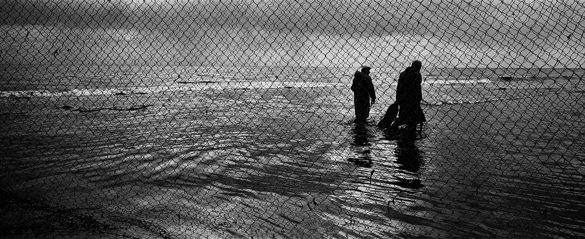Photogrphers image of two fishermen wading through calf-deep water on a beach. The image is taken through a fishing net.