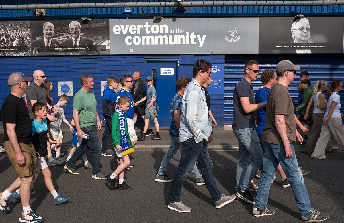 Photographer's image of a group of Everton football fans walking past a backdrop of the buildings painted in blue. A sign above reads "Everton in the Community".