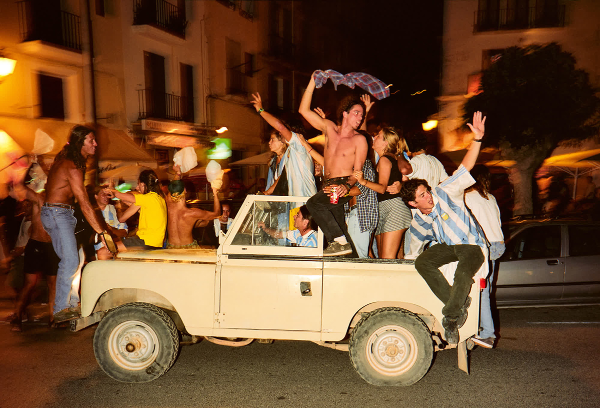 A nighttime streetscape with an open topped jeep and young people dancing on it.