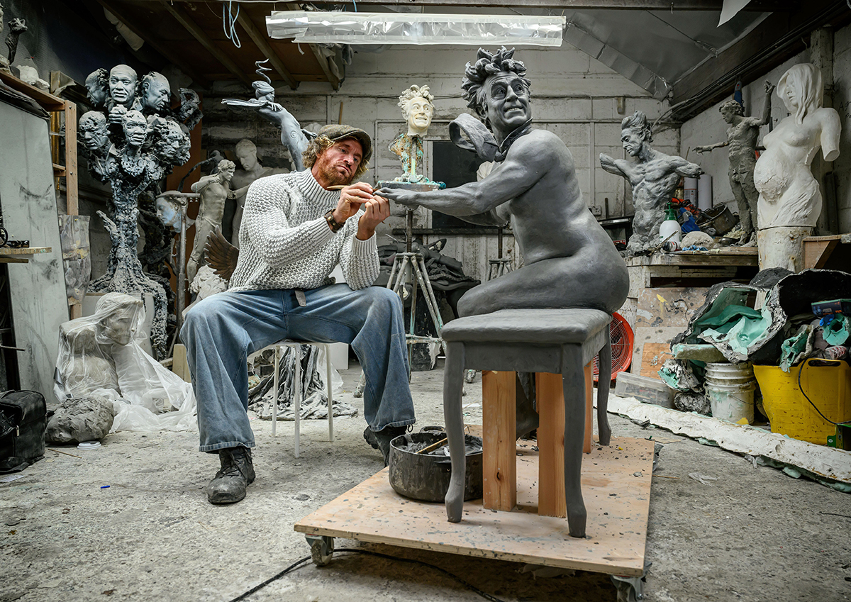 Sculptor working with piece., surrounded by many other pieces.
