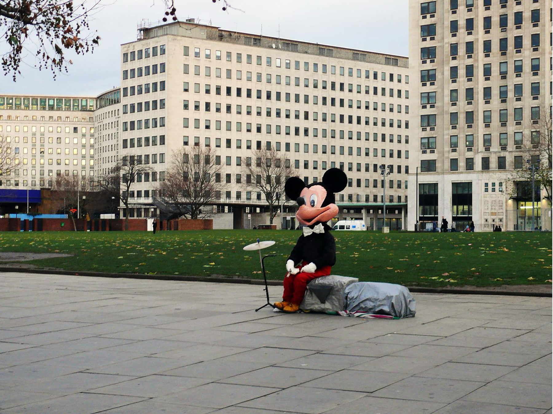 Photograph of a person dressed in a mickey mouse costume sat is a city scene alongside a cymbal.