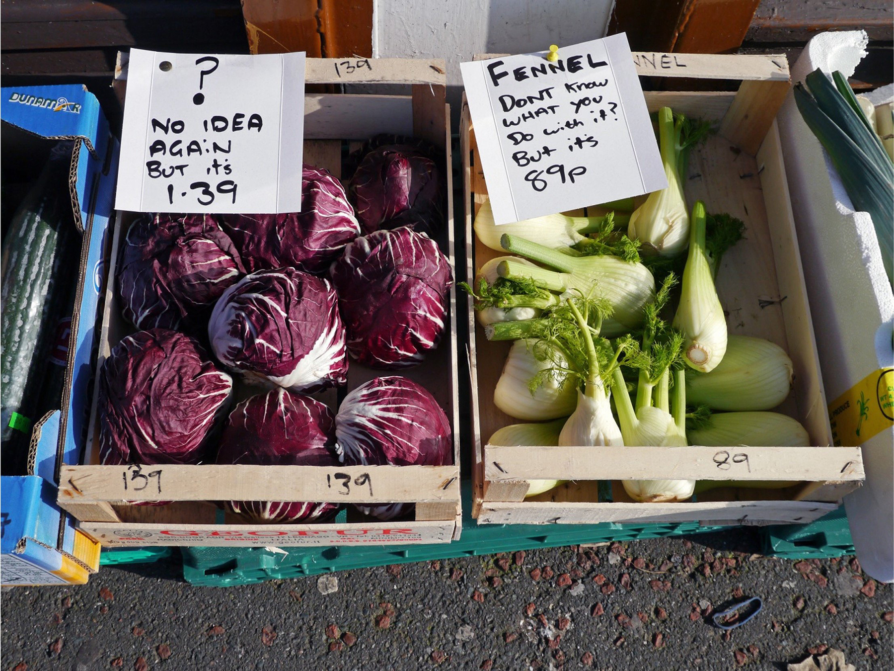Phoograph of two crates of vegatables with price signage that suggests that the vendor does not know what the vegetables are.